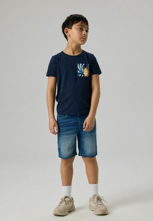 Chłopiec stojący, ubraný w granatowy t-shirt z kwiatową kieszenią, dżinsowe szorty, białe skarpetki i beżowe trampki, na tle jednolitego szarego tła.