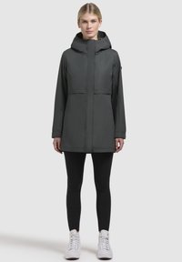 Donkergrijze waterproof jas met capuchon, met een rits aan de voorkant en twee zakken. Iets losse pasvorm, gecombineerd met zwarte leggings en witte sneakers.