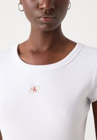 Bílý žebrovaný top s kulatým výstřihem a krátkými rukávy, s logem Calvin Klein v červené a béžové barvě blízko výstřihu.