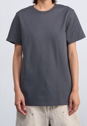 Osoba ubrana w jednokolorowy ciemnoszary krótki rękaw T-shirt i jasnobeżowe spodnie, stojąca na tle jasnoszarym.