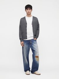 Jeune homme debout portant un cardigan gris boutonné sur un t-shirt blanc, un jean large déchiré avec des patchs, et des baskets blanches.