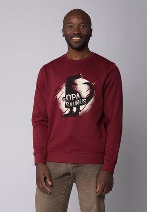 Burgundy sweatshirt with a black dinosaur graphic and "DPA SAURUS" text. Features ribbed cuffs and a crew neck. Fabric has a smooth texture.
