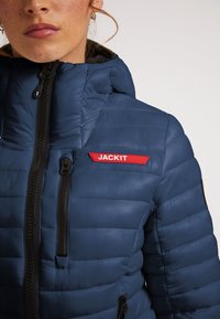 Blå pufferjakke med polstret overflade, udstyret med en høj krave, frontlynlås og et rødt logo-patch med teksten "JACK1T" på brystet.