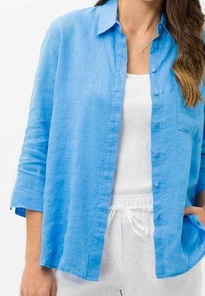 Woman wearing a light blue button-up shirt over a white tank top and white drawstring pants, with one hand in pocket.