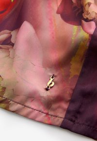 Satinė medžiaga rožinės ir violetinės spalvų tonais, puošta gėlių raštu. Krašte pritvirtinta mažytė auksinė pakabuko forma su raide „b“.