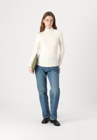 Kremfarget høyhalset genser kombinert med lyseblå jeans. Modellen holder en olivengrønn clutch; fottøyet er svart. Enkelt, figursydd design.