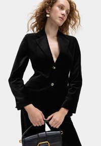 Czarna aksamitna marynarka z głębokim dekoltem w kształcie litery V, z pojedynczym złotym zapięciem i długimi rękawami. Modelka trzyma fakturowaną czarną torebkę.