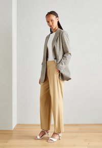 Karierter Blazer in Grau und Beige, kombiniert mit hellbraunen hochflorigen Hosen. Weiße Riemchen-Absatzsandalen runden das Outfit ab. Glatte Textur.