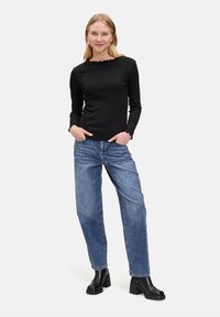 Zwarte lange mouw top met gegolfde halslijn, gecombineerd met lichtblauwe rechte jeans en zwarte chunky heeled laarzen. Witte achtergrond.