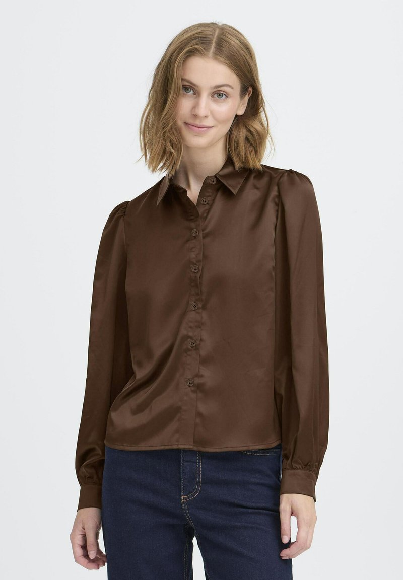 Blusa lunga in satin marrone con maniche lunghe, apertura a bottoni sul davanti, colletto a punta e dettagli arricciati sulle spalle. Abbinata a jeans in denim scuro.