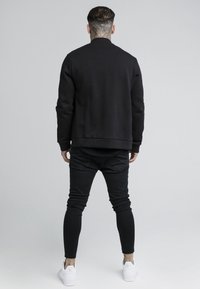 Felpa nera con scollatura rotonda e polsini a coste, abbinata a jeans skinny neri e sneakers bianche, vista da dietro.