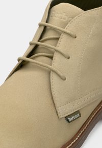 Botte chukka en daim beige avec quatre lacets, un design cousu, et une petite étiquette verte portant l'inscription « Barbour » sur le côté.