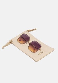 Sunglasses with a square shape, gradient purple lenses, and gold accents, resting on a light beige soft pouch marked "LIU•JO."