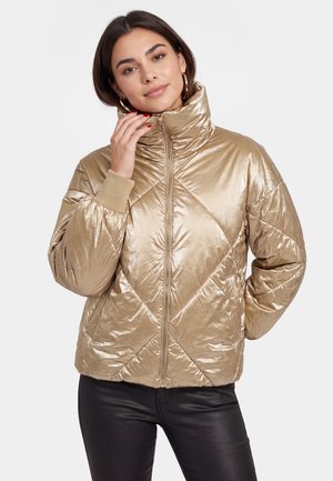 Frau mit dunklen Haaren, die eine goldene gesteppte Daunenjacke mit hohem Kragen und schwarze Hose trägt, stehend vor neutralem Hintergrund.