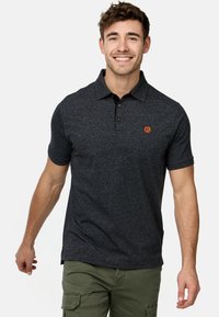 Polo shirt gris foncé à manches courtes, en tissu texturé, avec patte de boutonnage à trois boutons et logo orange discret sur la poitrine. Porté avec un pantalon cargo.
