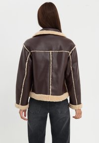 Brun kunstskinnjakke med beige fleecefôr og trim, med krage og kort design, synlige sømdetaljer.
