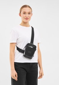 Borsa a tracolla nera realizzata con materiali durevoli, caratterizzata da una forma rettangolare, cinturino regolabile, tasca frontale con zip e dettaglio logo testuale.