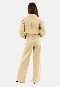 Conjunto de dos piezas en color beige que incluye una camisa con cuello y pliegue en la espalda, y pantalones de pierna ancha de talle alto con cinturilla elástica.