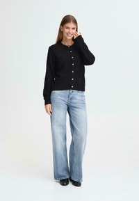 Svart strikket cardigan med rund hals, fem knapper foran, og lange ermer, sammen med lyseblå vide jeans.