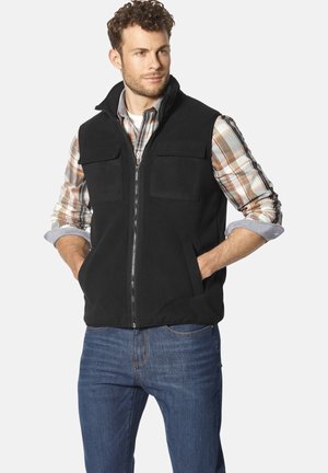 Black fleece vest with high collar, front zipper, and two chest pockets, worn over a plaid shirt and paired with blue jeans.