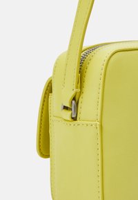 Borsa a tracolla gialla in pelle con forma rettangolare, compartimento con zip, cuciture a vista e dettaglio della fibbia in metallo argentato.