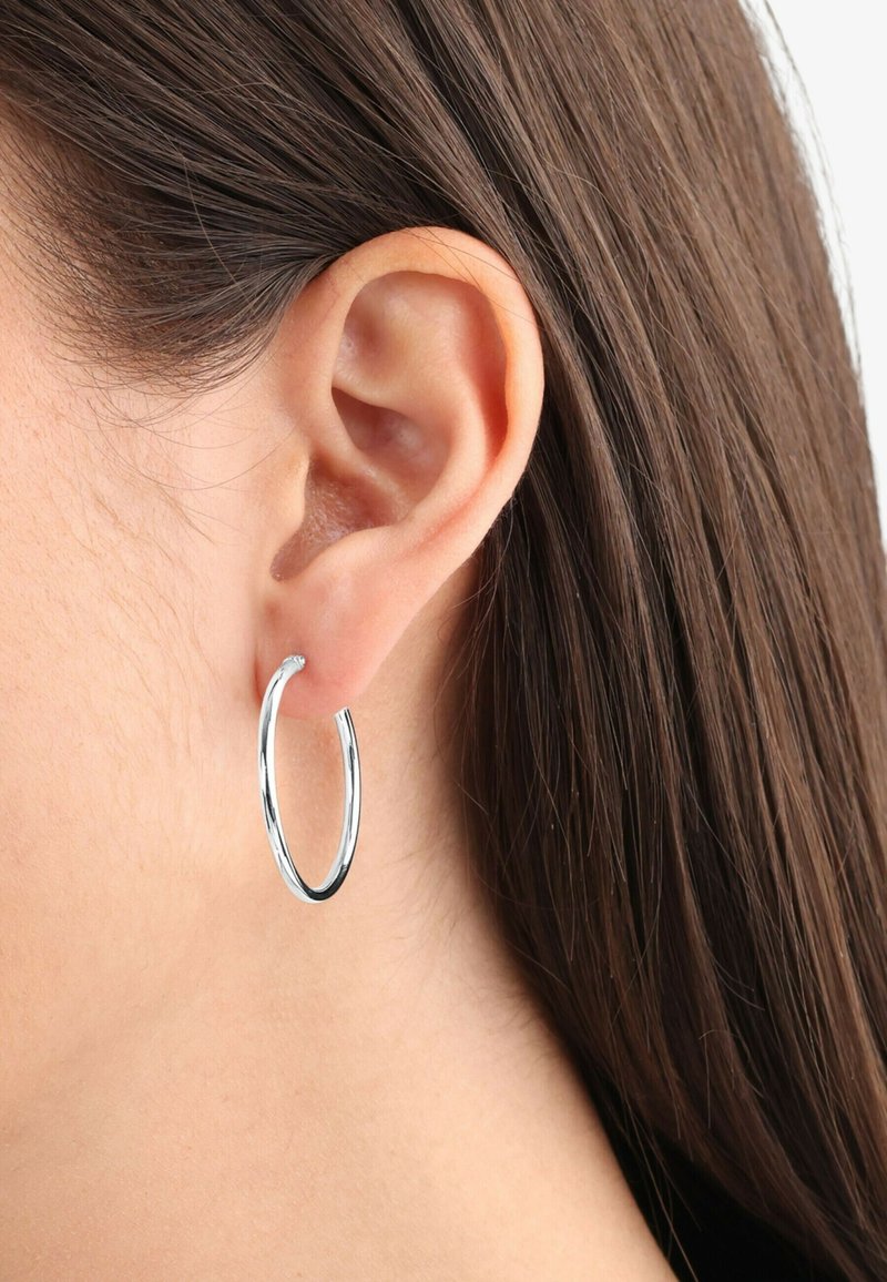 Pendiente de aro plateado colgando de una oreja, con una textura suave y brillante y un diseño circular, con un perfil delgado y minimalista.