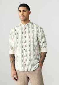Chemise à col crème avec des rayures verticales noires et des motifs géométriques, associée à un pantalon beige clair. Tissu texturé, coupe décontractée.