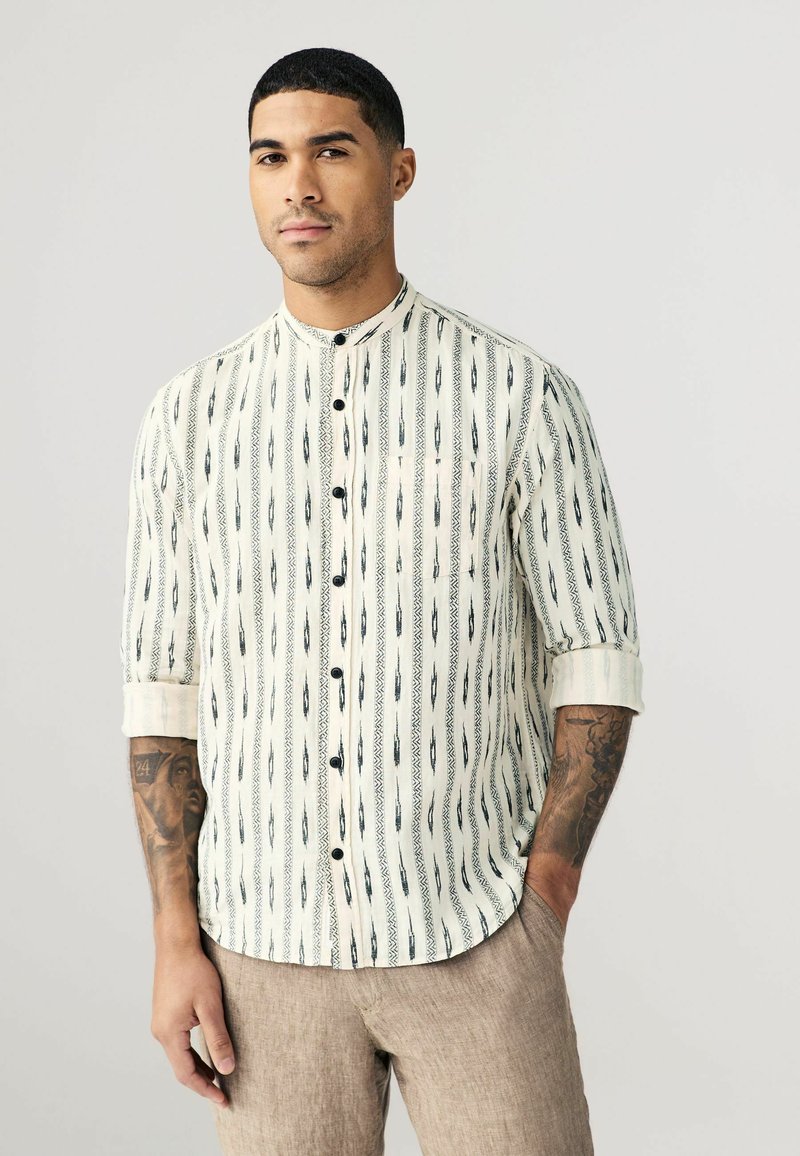 Chemise à col crème avec des rayures verticales noires et des motifs géométriques, associée à un pantalon beige clair. Tissu texturé, coupe décontractée.