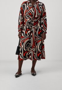 Vestido envolvente impreso con patrones en espiral en negro, rojo y beige; mangas largas y detalle de cintura con lazo. Combinado con zapatos planos negros y un bolso.