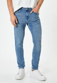 Vaqueros de mezclilla de color azul claro con un corte ajustado, diseño de cinco bolsillos y pasadores para cinturón. Lleva puestos con zapatillas blancas y una camiseta blanca sencilla.