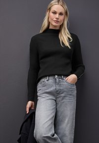Czarny sweter z prążkowanej dzianiny o wysokim kołnierzu zestawiony z jasnoszarymi spodniami o szerokich nogawkach. Model trzyma ciemną kurtkę. Gładkie tło.