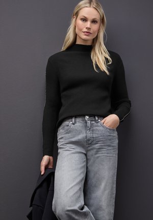 Zwarte geribbelde gebreide trui met hoge hals, gecombineerd met lichtgrijze wijde jeans. Model houdt een donkere jas vast. Eenvoudige achtergrond.