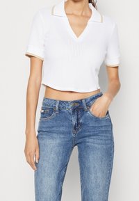 Top court polo blanc à rayures, avec un col en V et des manches courtes, arborant une bordure beige aux poignets et au col. Assorti avec un jeans en denim bleu.