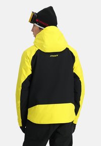 Veste de ski noire et jaune vif avec une capuche, arborant un design élégant, une texture lisse et le logo Spyder au dos.