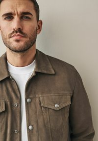 Bruine suède jas met een knoopsluiting aan de voorkant, twee borstzakken en zilveren hardware. Gemaakt over een wit shirt tegen een neutrale achtergrond.