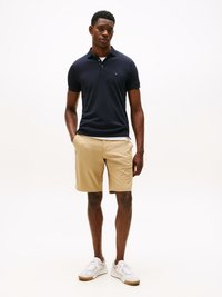 Marineblå polo med korte ærmer, to knapper ved kraven, kombinert med khaki shorts. Hvite sneakers med teksturert såle fullfører stilen.