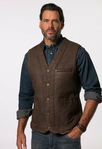 Gilet en cuir marron avec fermeture à boutons, présentant une poche poitrine et deux poches avant. Porté par-dessus une chemise en denim à manches retroussées.