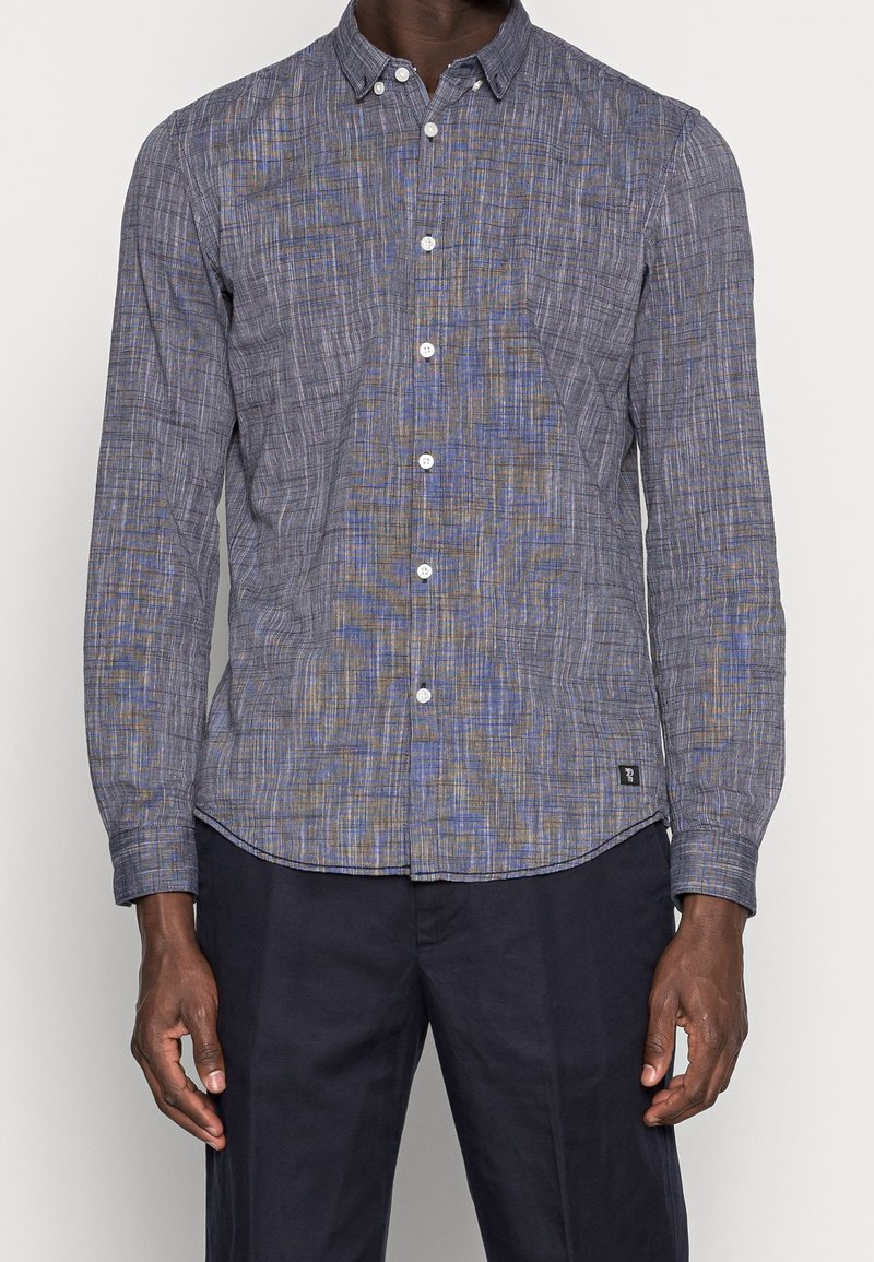 Homme portant une chemise à manches longues boutonnée à motif à carreaux gris et bleus et un pantalon foncé, debout devant un fond uni.