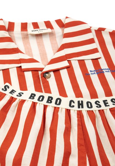 Chemise à rayures verticales rouges et blanches avec col, un bouton, étiquette « Bobo Choses » et texte brodé indiquant « I'm not going out Club. »