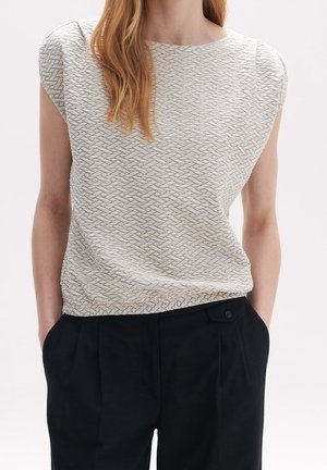Un mannequin porte un haut blanc texturé à manches courtes, aux épaules structurées, ainsi qu’un pantalon noir plissé, les mains dans les poches.