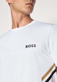 Hvit atletisk skjorte laget av glatt stoff, med prikkete mønster, "BOSS"-logo i svart, og diagonale stripete detaljer i gull og svart.