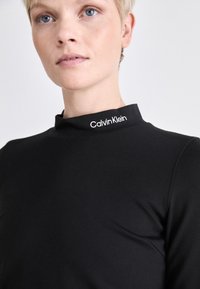 Haut noir à col montant en tissu lisse, arborant un logo Calvin Klein blanc sur le col. Design simple et ajusté.