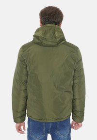 Giacca puffer con cappuccio di colore verde oliva, dalla texture liscia, con polsini elasticizzati e design a lunghezza vita, abbinata a jeans blu.