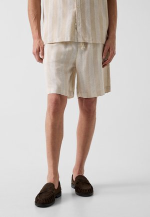 Uomo che indossa pantaloncini beige a righe e camicia abbottonata coordinata con mocassini marrone scuro su uno sfondo chiaro semplice.