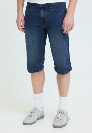 Homme portant un short en denim bleu foncé arrivant aux genoux, des chaussettes blanches et des baskets gris clair, debout sur un fond clair uni.