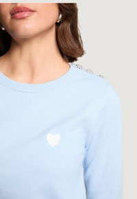 Jasnoniebieski sweter z okrągłym dekoltem, ozdobiony małą białą naszywką w kształcie serca oraz dekoracyjnymi akcentami na ramieniu. Miękka dzianinowa faktura.