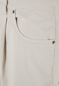 Bēšas kokvilnas bikses ar priekšējo kabatu. Tām ir neliela tekstūra, šūtas šuves un metāla pogas detaļa.