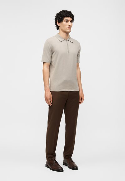 Άνδρας που φοράει μπεζ κοντομάνικο πόλο με φερμουάρ, καφέ παντελόνια και σκούρα καφέ επίσημα παπούτσια, μπροστά σε απλό φωτεινό φόντο.