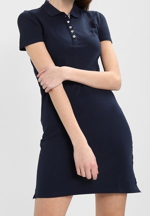 Woman wearing a fitted navy blue short-sleeve polo dress with buttons down the chest, arms crossed in front of her body.