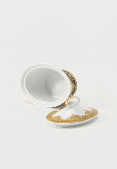 Λευκό πορσελάνινο βάζο με χρυσά διακοσμητικά μοτίβα, που βρίσκεται ξαπλωμένο στο πλάι με το καπάκι που ταιριάζει δίπλα του σε λευκή επιφάνεια.